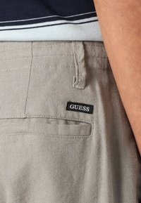Bež lanene hlače vsebujejo zadnji žep z black oznako blagovne znamke "GUESS". Tkanina ima teksturiran finish in je delno vidna nad mornarsko modro srajco.