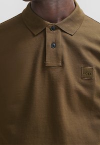 Polo marron en tissu texturé, doté d'un col classique, de deux boutons noirs et d'un petit logo embossé sur le côté gauche.