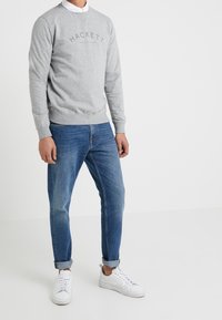 Sudadera gris con cuello redondo y logotipo "HACKETT" en relieve, combinada con jeans azules y zapatillas blancas. Ajuste casual y textura de la tela visible.