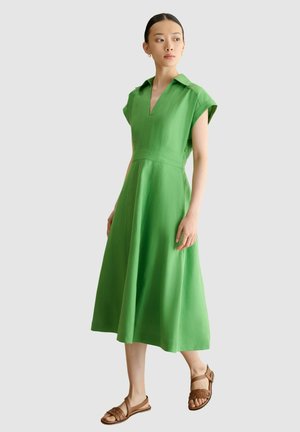 Mujer con un vestido midi verde con mangas farol y sandalias planas marrones, de pie frente a un fondo liso de color gris claro.