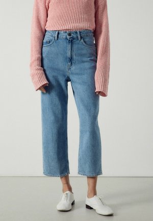 Persona che indossa jeans cropped a vita alta azzurri chiari, un maglione lavorato a maglia rosa con maniche lunghe e scarpe bianche con lacci, in piedi davanti a uno sfondo semplice.