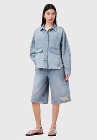 Giacca oversize in denim azzurro chiaro con due tasche frontali e orlo sfrangiato, abbinata a pantaloni corti abbinati fino al ginocchio con un dettaglio strappato.