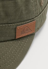 Groene stoffen pet met een gestructureerde afwerking, voorzien van een bruine leren patch met logo, en contrasterende stiksel langs de rand.