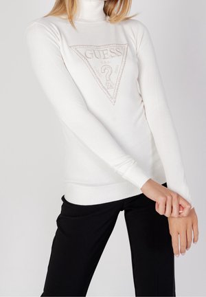Kvinde iført hvid GUESS højhalset sweater med logo og sorte bukser, justerer ærmet og står mod en ensfarvet lys baggrund.