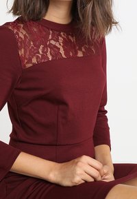 Bordowa sukienka z dopasowanym gorsetem, długimi rękawami i przezroczystą koronkową wstawką przy dekolcie. Gładki materiał o subtelnej teksturze.