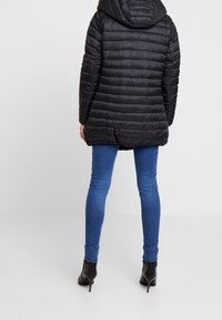 Chaqueta acolchada negra con capucha, que presenta una textura acolchada y una longitud que llega hasta la mitad del muslo, combinada con jeans ajustados azules y botas negras hasta el tobillo.