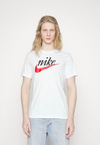 T-shirt bianco in cotone con un grande logo Nike nero e rosso sul petto. Maniche corte; design standard con colletto a girocollo.