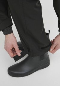 Sorte vanntette bukser med justerbare velcro mansjetter, brukt over svarte gummiankelsko, vist mens mansjettene strammes. Glatt tekstur.