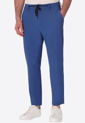 Uomo che indossa pantaloni slim-fit blu con coulisse nera e sneakers bianche, in piedi con una mano in tasca su sfondo bianco.
