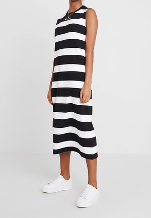 Kobieta ubrana w bezrękawnikową, czarno-białą sukienkę midi w poziome paski, połączoną z białymi trampkami na jednolitym tle.