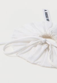 Λευκό υφασμάτινο scrunchie με υφή στην επιφάνεια και πιεσμένο σχέδιο, που διαθέτει ελαστική ταινία και μια μικρή ετικέτα με λογότυπο.