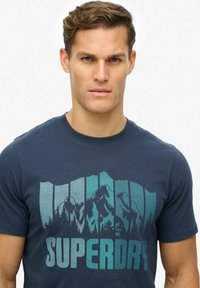 Granatowa bawełniana koszulka z grafiką przedstawiającą góry i drzewa, z wyrazem "SUPERDRY" napisanym dużą, gradientową czcionką.