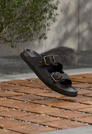 Mørkebrune vevde skinnsandaler med to gullspenner som svever over trefliser utendørs.