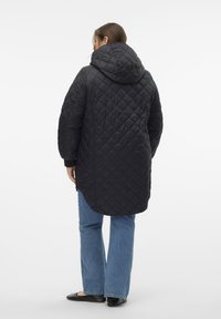 Abrigo largo acolchado negro con capucha, con un patrón de diamantes, textura suave y puños acanalados, combinado con jeans de campana en azul claro.