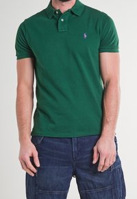 Grön polo skjorta i texturerad tyg med en två-knappsknäppning, med en liten lila logotyp på vänster bröst.