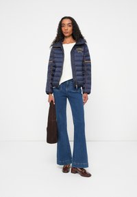 Marineblå pufferjakke med kviltede sektioner og orange piping, hvid top, flagrende blå jeans og brun håndtaske. Brune fodtøj med hardware-detaljer.