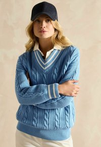 Pulover tricotat de culoare albastru deschis, cu modele cu torsade și texturate, având un guler și manșete cu dungi albe, purtat peste o cămașă cu guler.