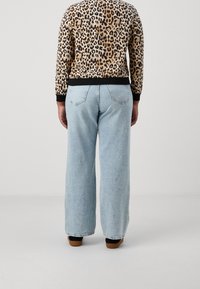 Personne portant un haut à manches longues imprimé léopard et un jean large bleu clair, debout de dos sur un fond uni clair.