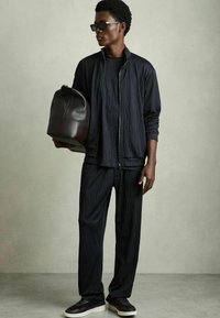 Veste zippée noire texturée, pantalon assorti et baskets foncées, accompagnés d'un sac à dos en cuir brun, le tout sur un fond neutre.