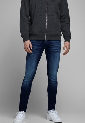 Mann trägt dunkelgrauen Hoodie mit Reißverschluss, eng anliegende blaue Jeans und weiße Sneaker, steht vor schlichtem grauem Hintergrund.