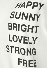 T-shirt in cotone bianco con testo stampato in nero in grassetto, con parole positive disposte verticalmente: "FELICE," "SOLEGGIATO," "BRILLANTE," "AMOREVOLE," "FORTE," "LIBERO."