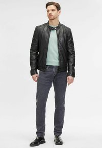 Giacca di pelle nera con colletto rialzato, indossata sopra una camicia azzurrino chiaro, abbinata a jeans grigi e scarpe nere. Design casual e aderente.