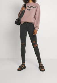 Sudadera rosa con el texto "MOVIMIENTO", jeans skinny negros rasgados y sandalias negras con doble tira. Bolsa de hombro negra sencilla.