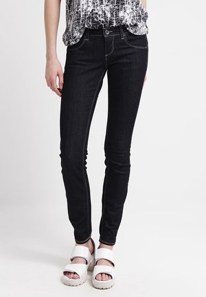 Schmal geschnittene schwarze Jeans, kombiniert mit einem schwarz-weiß gemusterten Oberteil und weißen Plateausandalen mit zwei breiten Riemen.