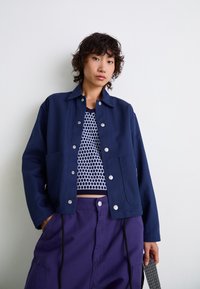 Veste bleu marine avec une fermeture à bouton et deux grandes poches avant, portée par-dessus un haut en maille à motif. Pantalons violets avec une taille élastique.