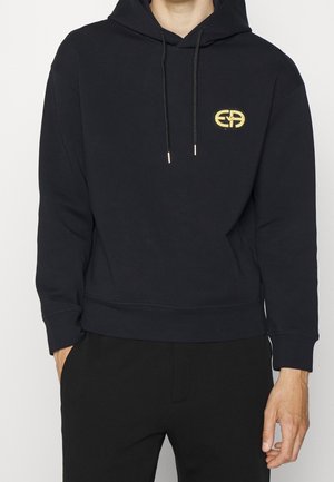 Mand iført sort hættetrøje med gyldent broderet logo på venstre bryst og sorte bukser, hænder afslappet ved siderne.