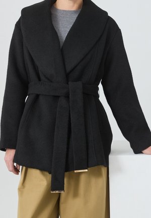Personne portant un manteau en laine noir ceinturé sur un pull gris, associé à un pantalon beige, avec une main posée sur une surface blanche.
