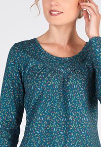 Türkise Bluse mit langen Ärmeln und floralem Muster in Rot, Grün und Blau. Sie verfügt über einen rundem Halsausschnitt und geraffte Details an der Brust.