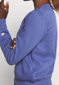 Lila sweatshirt tillverkad av mjukt material, med rundad halsringning och ribbade ärmslut. En broderad logotyp är synlig på vänster ärm.