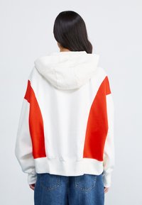 Sweat-shirt à capuche blanc avec des accents rouges, présentant une coupe décontractée, de longues manches et des poignets côtelés. Texture douce, sans logos ni motifs visibles.