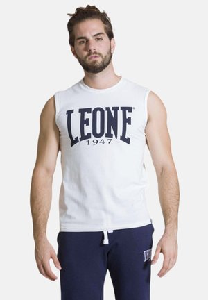 Man met baard en samengebonden haar, die een wit mouwloos shirt en donkerblauwe joggingbroek draagt, staand met het gezicht naar voren tegen een effen achtergrond.