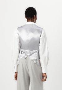Gilet en satin argenté avec une texture lisse, associé à une chemise blanche à manches bouffantes et un pantalon gris, vue de dos.