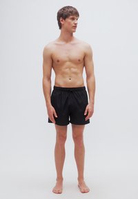 Μαύρα μαγιό ανδρικά με ελαστική μέση και κορδόνι, φορεμένα από έναν άνδρα που στέκεται μπροστά σε ένα απλό φωτεινό φόντο.