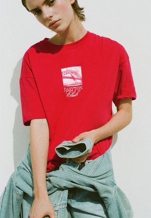 Giovane persona con t-shirt rossa "Daryton Club" che tiene una giacca di jeans piegata davanti a uno sfondo chiaro e uniforme.