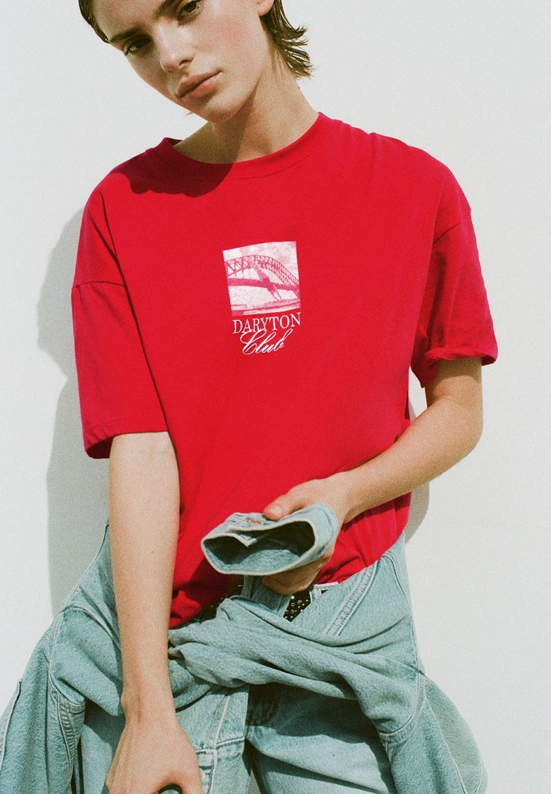 Jeune personne portant un t-shirt rouge « Daryton Club » tenant une veste en jean pliée sur un fond clair uni.