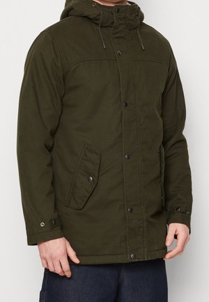 Mand iført en olivengrøn hættejakke med knapper foran og klaplommer, kombineret med mørkeblå bukser.