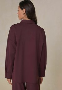 Burgundy oversized shirt featuring a collared neckline, buttoned cuffs, and a smooth texture. Back view highlights a clean seam design.