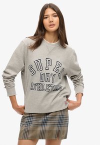 Sweat-shirt gris avec un texte audacieux en bleu marine indiquant "SUPER DRY ATHLETE." Assorti à une mini-jupe à carreaux aux tons bleus et beiges.