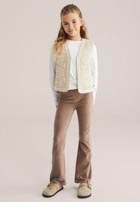 Gilet beige ricamato sopra una maglietta bianca, abbinato a pantaloni a zampa d'elefante marroni a coste e scarpe slip-on chiare. Dettagli semplici e testurizzati.