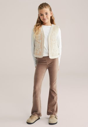 Beige geborduurd vest over een witte top, gecombineerd met bruine geribde flared broeken en lichtgekleurde instap schoenen. Eenvoudige, gestructureerde details.