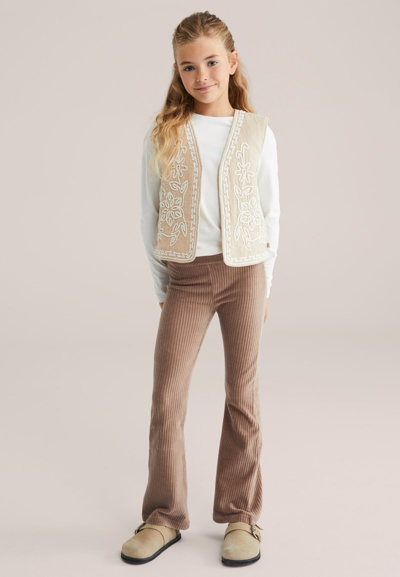 Gilet beige ricamato sopra una maglietta bianca, abbinato a pantaloni a zampa d'elefante marroni a coste e scarpe slip-on chiare. Dettagli semplici e testurizzati.