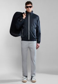 Chaqueta ligera navy con cierre frontal, puños elásticos y dos bolsillos laterales. Pantalones grises y zapatillas blancas; modelo llevando una bolsa negra.