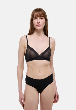 Femme aux cheveux bruns lisses portant un soutien-gorge noir transparent et une culotte taille haute noire, debout devant un fond blanc uni.