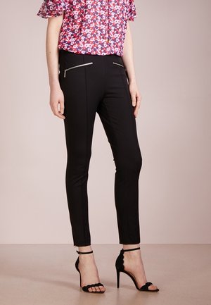 Vrouw draagt zwarte slim-fit broek met ritszakken aan de voorkant, gecombineerd met zwarte sandalen met hoge hak en een rode blouse met bloemenprint.