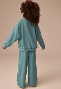 Maglione turchese a coste con maniche larghe abbinato a pantaloni a gamba larga coordinati. Il tessuto ha una consistenza morbida. Modello mostrato di spalle.