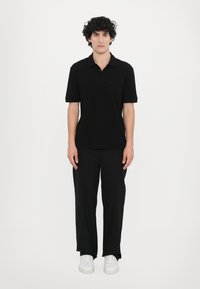 Svart polo skjorte med korte ermer, laget av myk stoff, kombinert med løstsittende svarte bukser og hvite joggesko i et minimalistisk miljø.